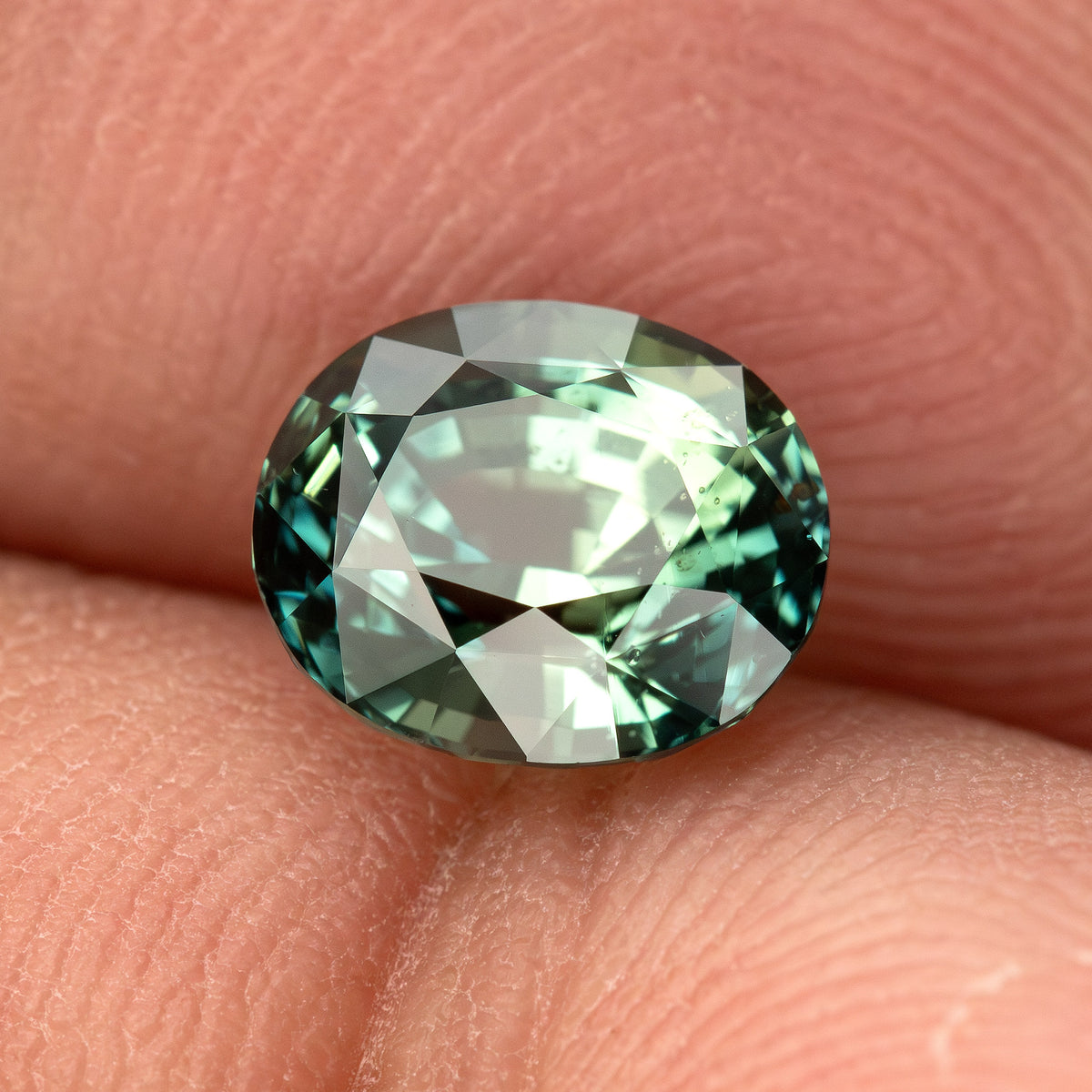 Oval teal blue-green sapphire, face-up view, resting on fingertips, showcasing its facets and color.