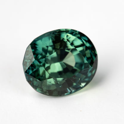 A face-up view of an oval teal-blue green natural sapphire with intricate facets on a white background.