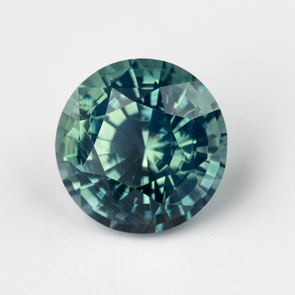 Teal blue-green round step-cut sapphire face-up on a light surface. View 1.
