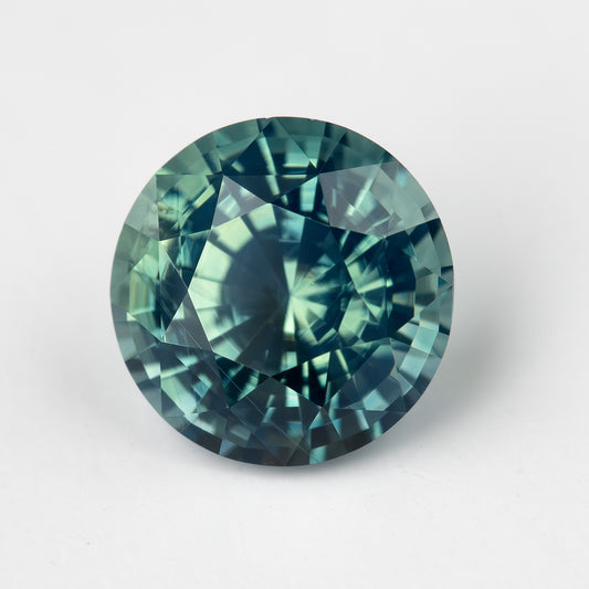 Teal blue-green round step-cut sapphire face-up on a light surface. View 1.