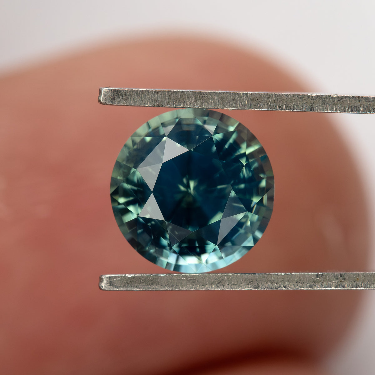 Teal blue-green round step-cut sapphire held in tweezers against a finger. View 6.