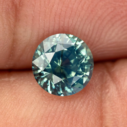 Teal blue-green round step-cut sapphire resting on fingertips for scale. View 3.