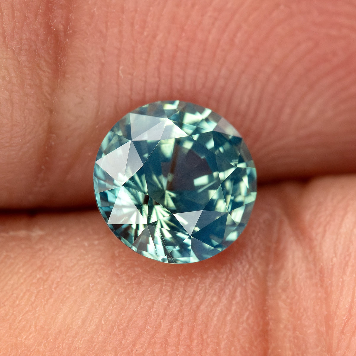 Teal blue-green round step-cut sapphire resting on fingertips for scale. View 20.
