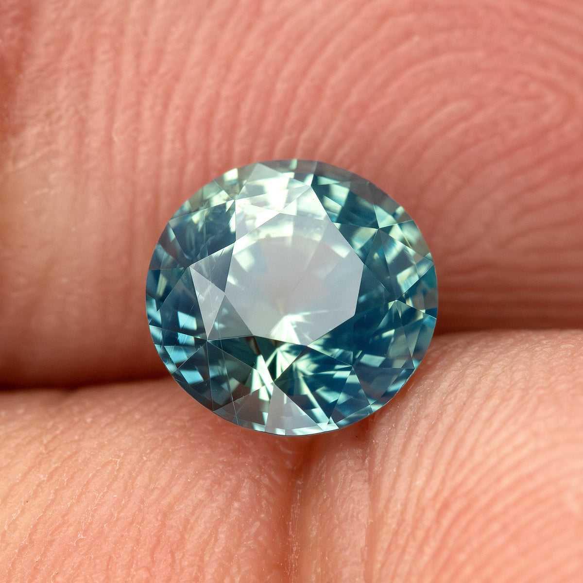 Teal blue-green round step-cut sapphire resting on fingertips for scale. View 4.