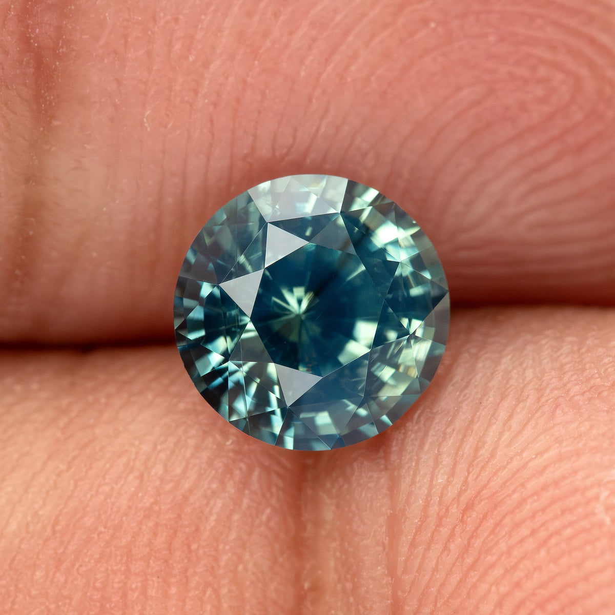 Teal blue-green round step-cut sapphire resting on fingertips for scale. View 5.