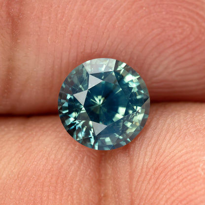 Teal blue-green round step-cut sapphire resting on fingertips for scale. View 5.
