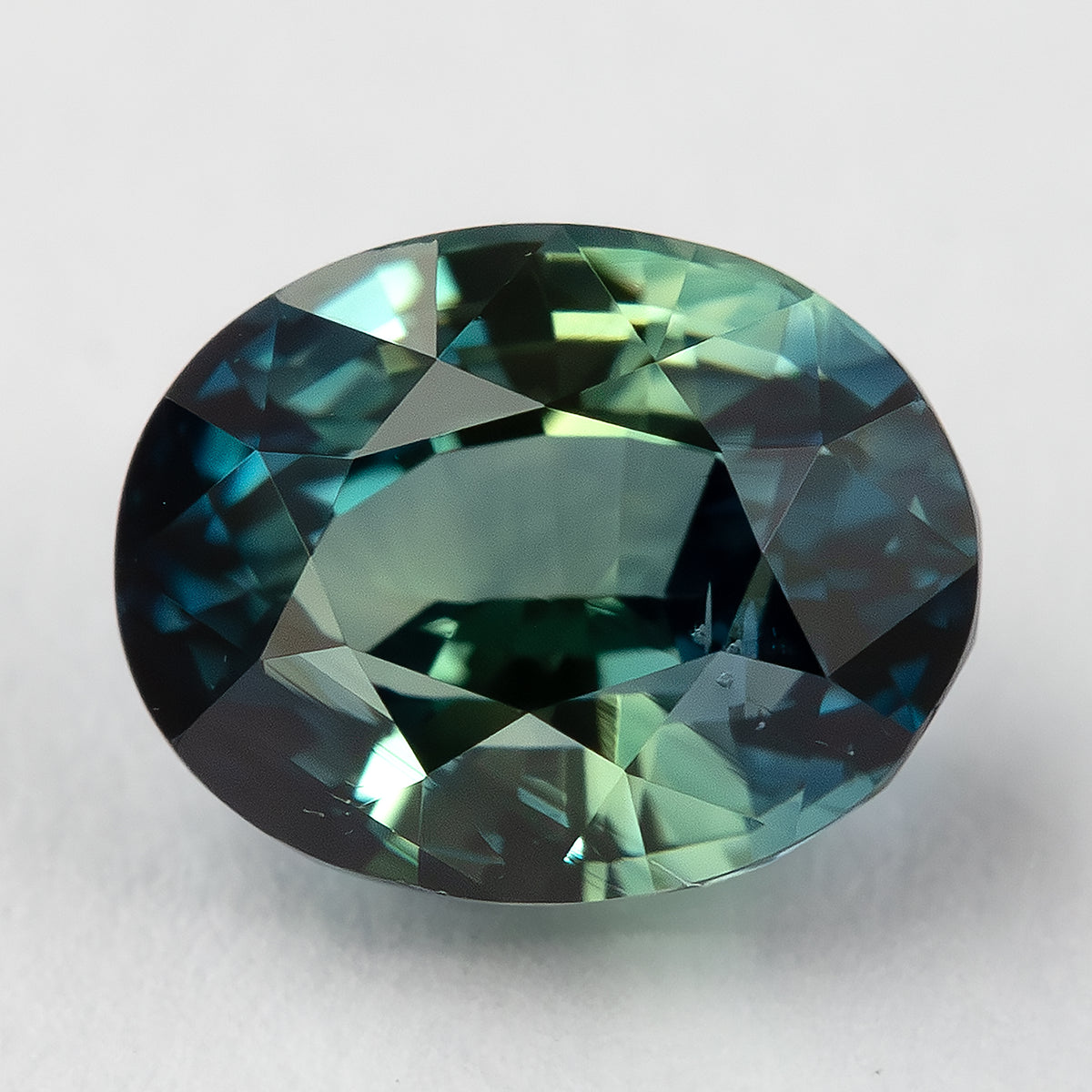 Teal blue-green oval sapphire lying face-up on a pale surface, showing its table and surrounding facets.
