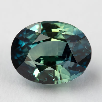 Teal blue-green oval sapphire lying face-up on a pale surface, showing its table and surrounding facets.
