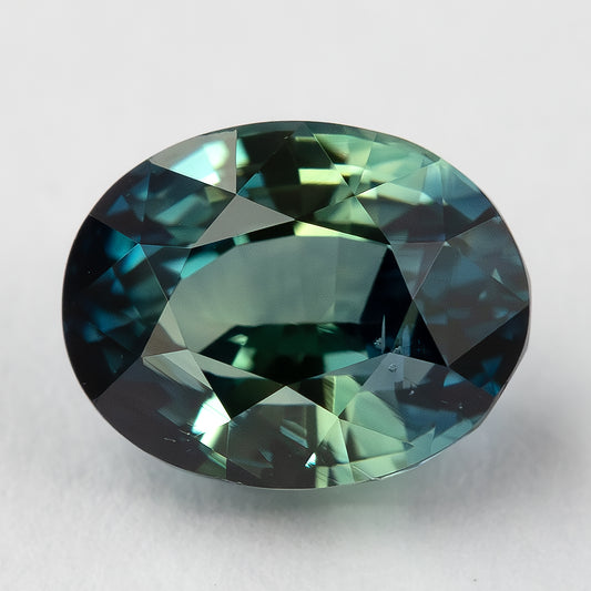 Teal blue-green oval sapphire lying face-up on a pale surface, showing its table and surrounding facets.