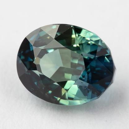 Oval parti sapphire tilted face-up on a pale surface, with blue and green reflections.