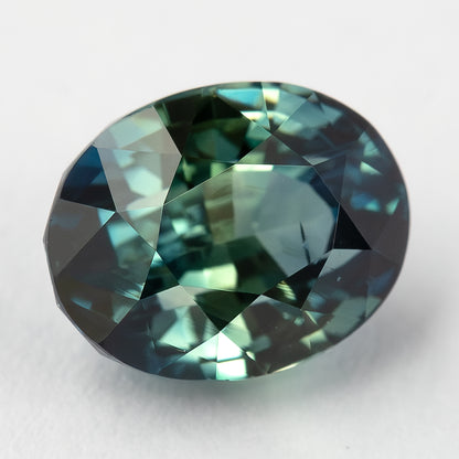 Face-up view of teal blue-green oval sapphire on a pale surface from an alternate angle.