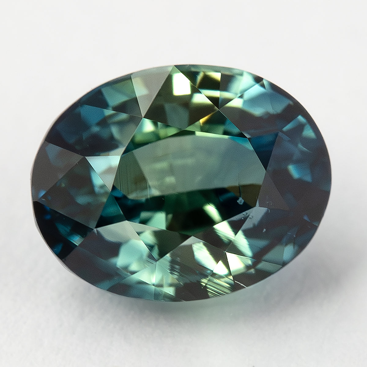 Oval sapphire lying face-up on a pale surface, showing broad facet reflections.