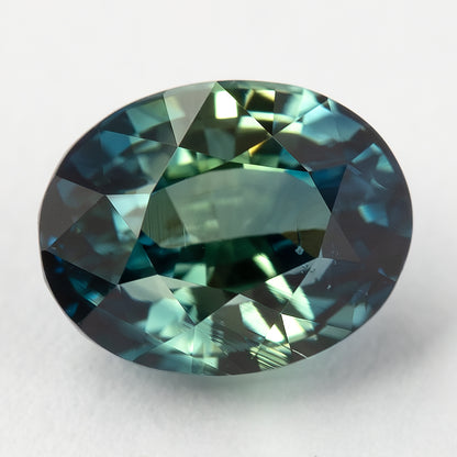 Oval sapphire lying face-up on a pale surface, showing broad facet reflections.