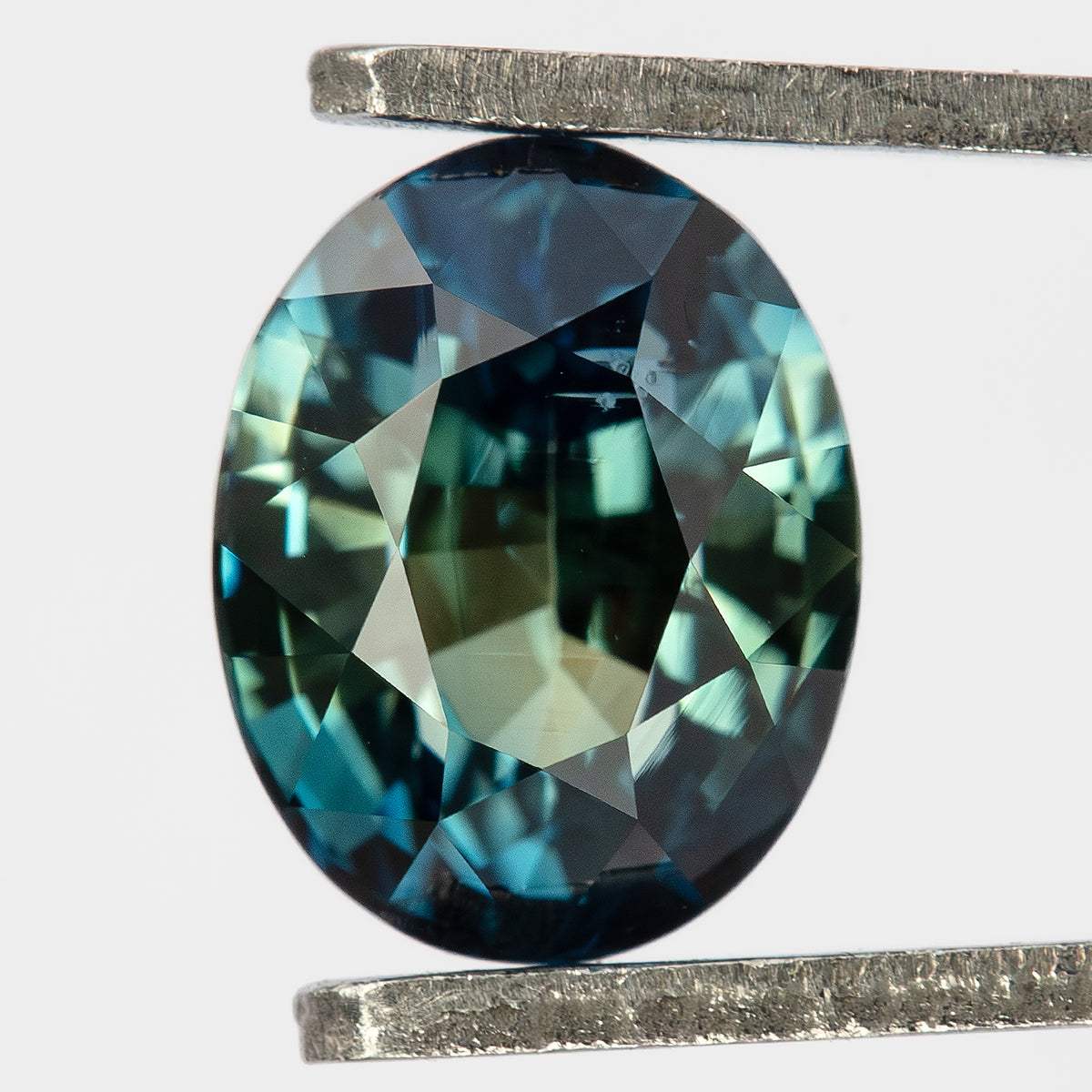 Teal blue-green oval sapphire held face-up vertically in metal tweezers.