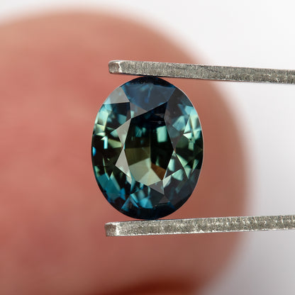 Face-up sapphire in tweezers against a blurred finger background.