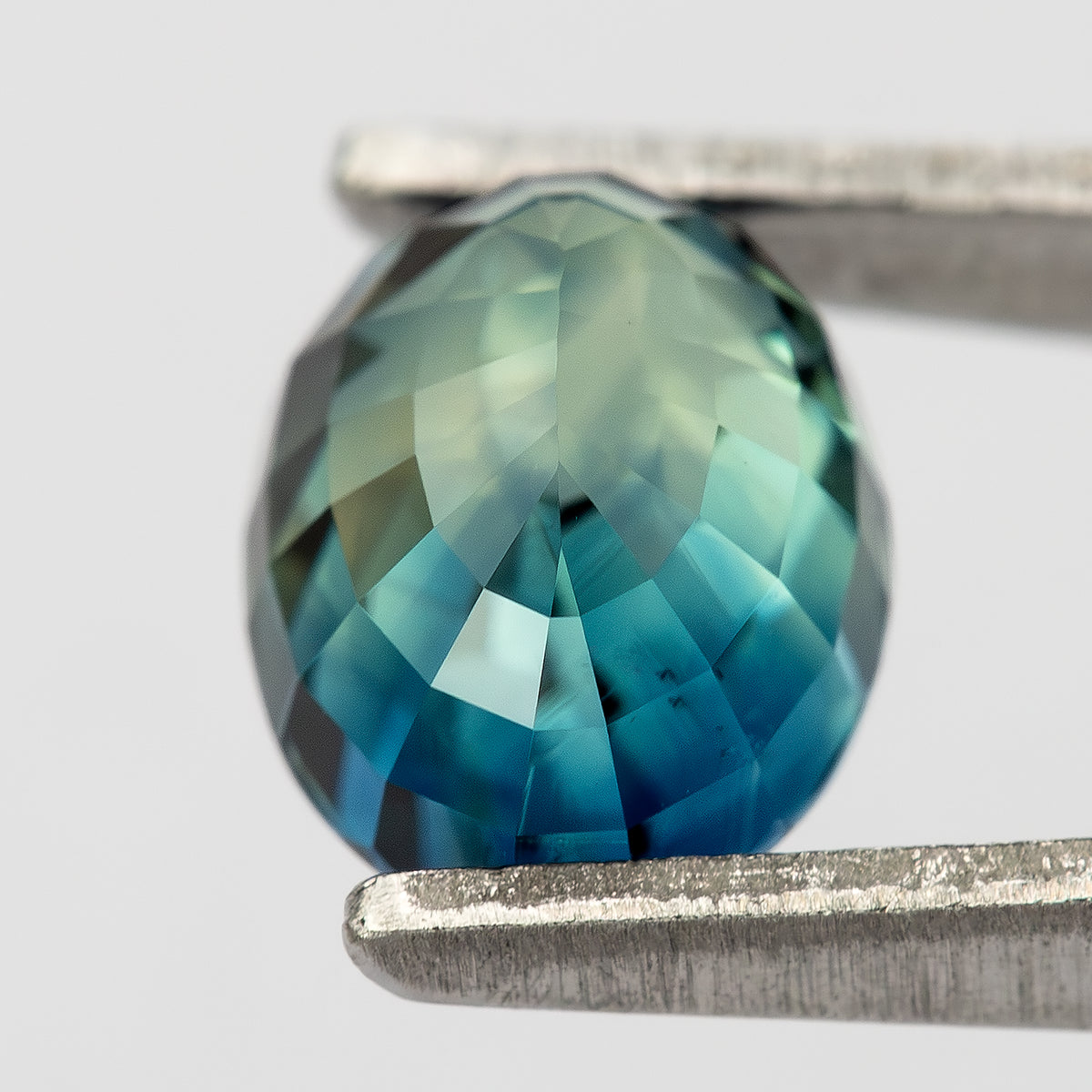 Tilted reverse view of sapphire in tweezers, showing blue-green pavilion facets.