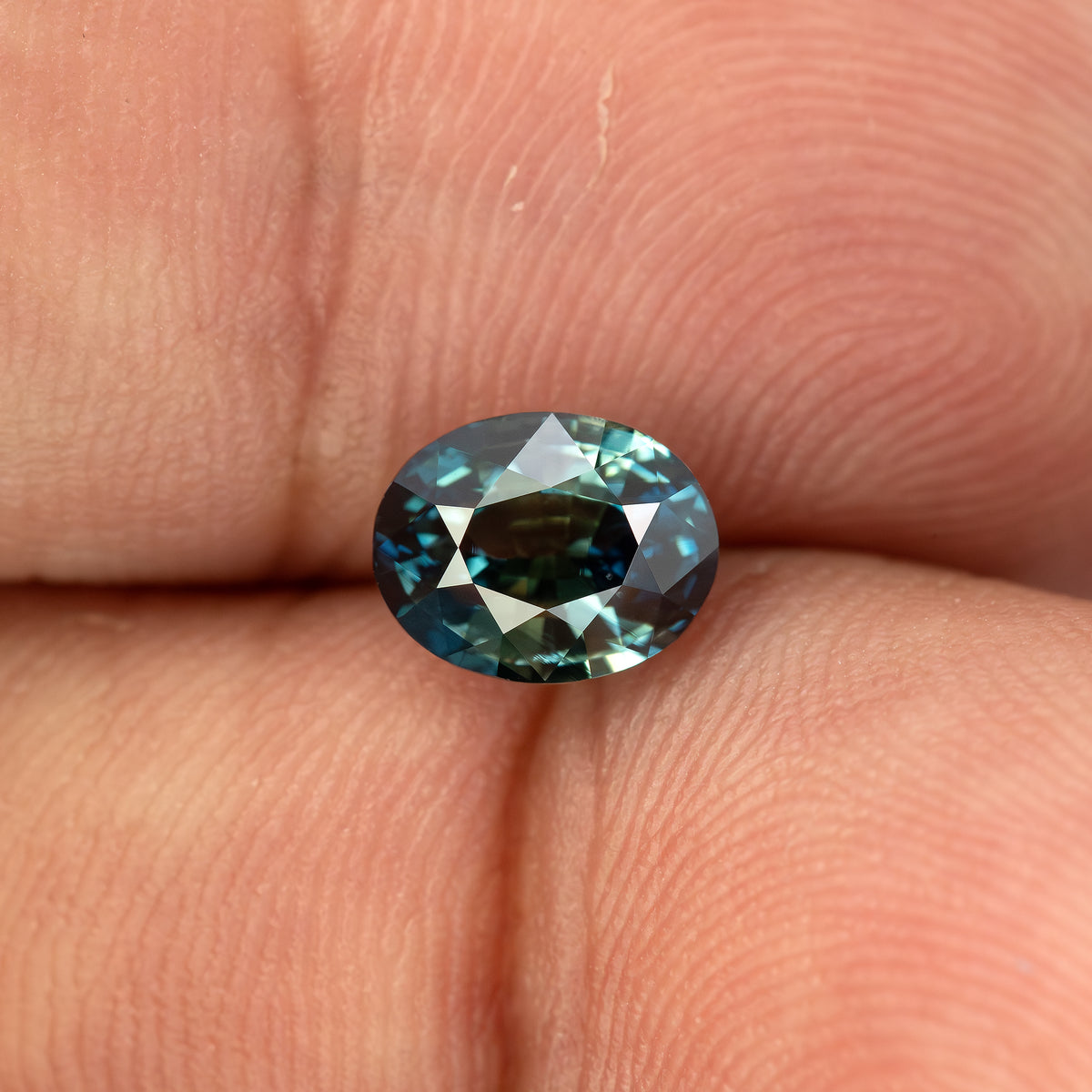 Teal blue-green oval sapphire resting face-up between fingertips for scale.