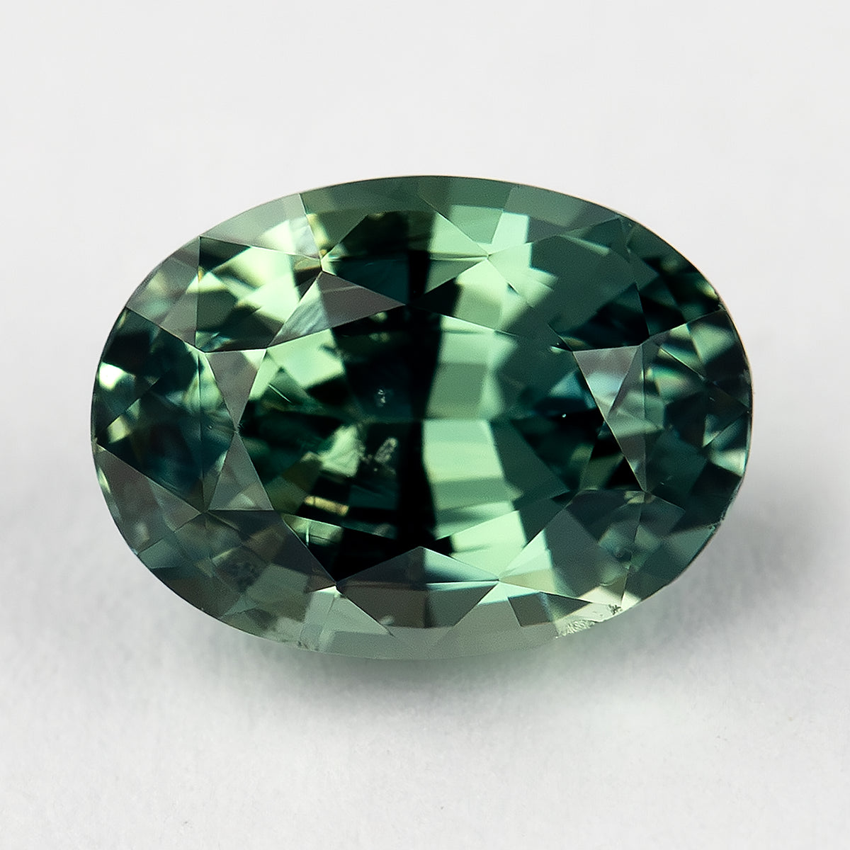 Teal-green oval natural sapphire face-up on a pale surface, with a dark central band and bright yellow-green facet reflections.