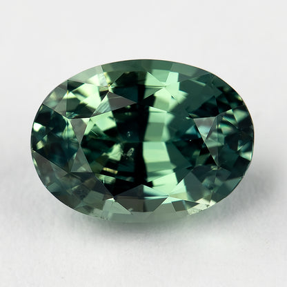 Teal-green oval natural sapphire face-up on a pale surface, with a dark central band and bright yellow-green facet reflections.