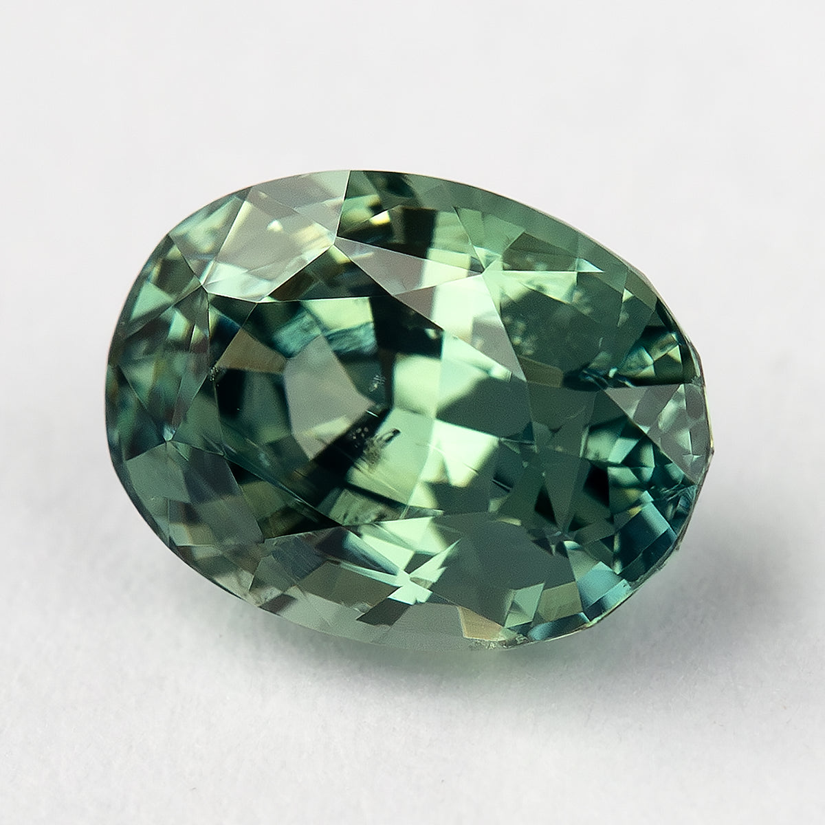 Teal-green oval sapphire tilted on a pale surface, showing the crown and bright angular reflections.