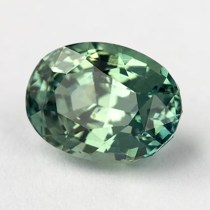 Tilted teal-green oval sapphire on a pale surface, with pale reflections across the center.