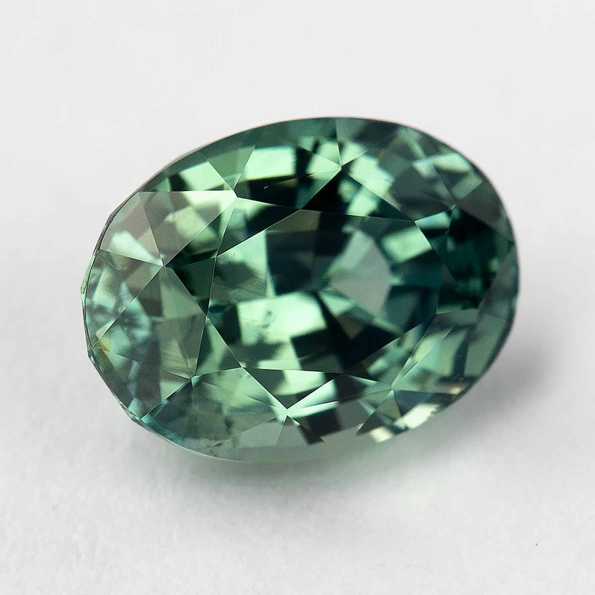 Angled face-up view of the teal-green oval sapphire, showing mixed green reflections across the crown.