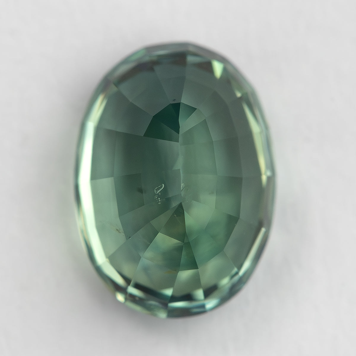 Back of the teal-green oval sapphire resting vertically on a pale surface, showing its pavilion facets.