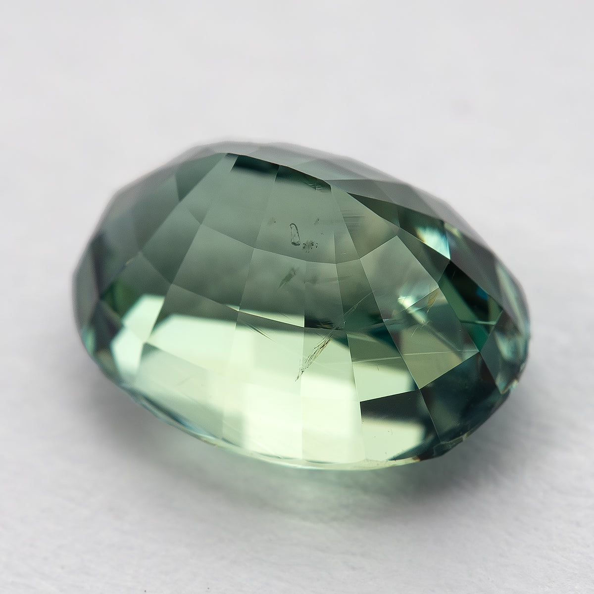 Teal-green oval sapphire tilted toward its back, showing the raised pavilion on a pale surface.