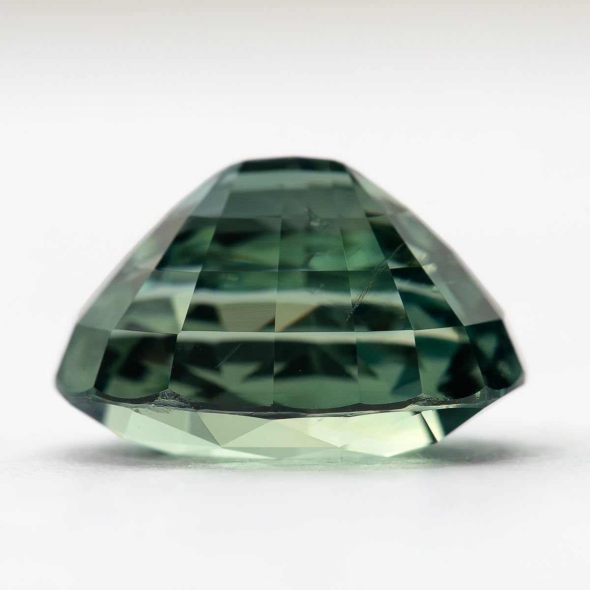 Side profile of the teal-green oval sapphire resting on a pale surface, showing crown height and pavilion depth.