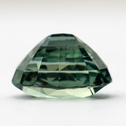 Side profile of the teal-green oval sapphire resting on a pale surface, showing crown height and pavilion depth.