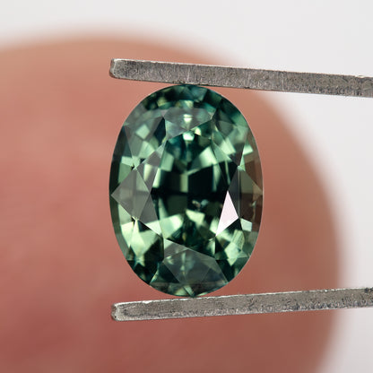 Teal-green oval sapphire face-up in tweezers against a fingertip for scale.