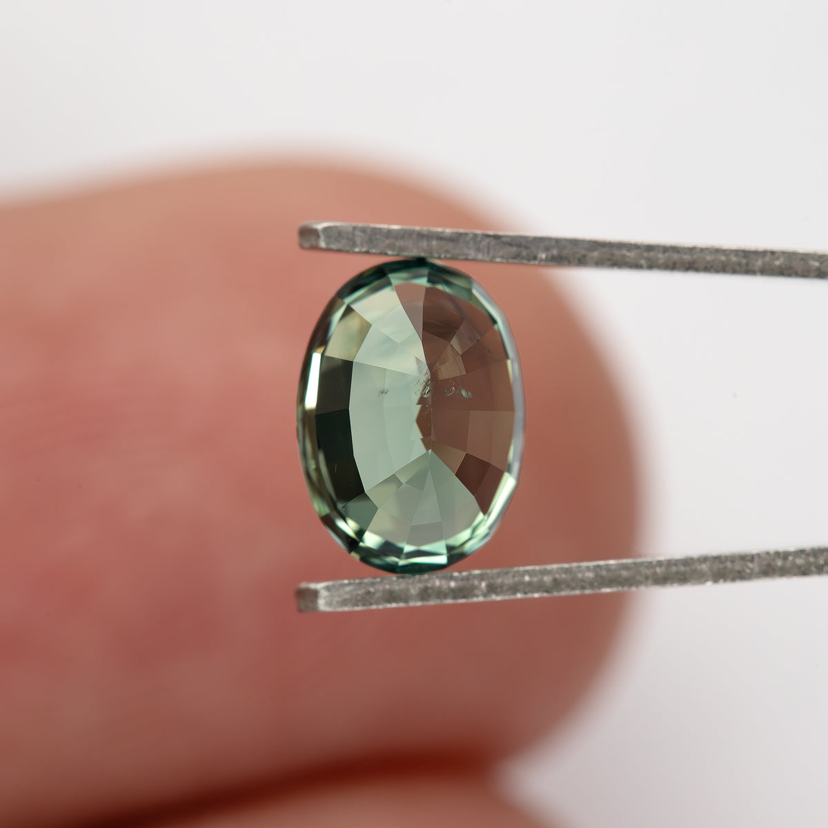 Teal-green oval sapphire turned backward in tweezers against a fingertip.