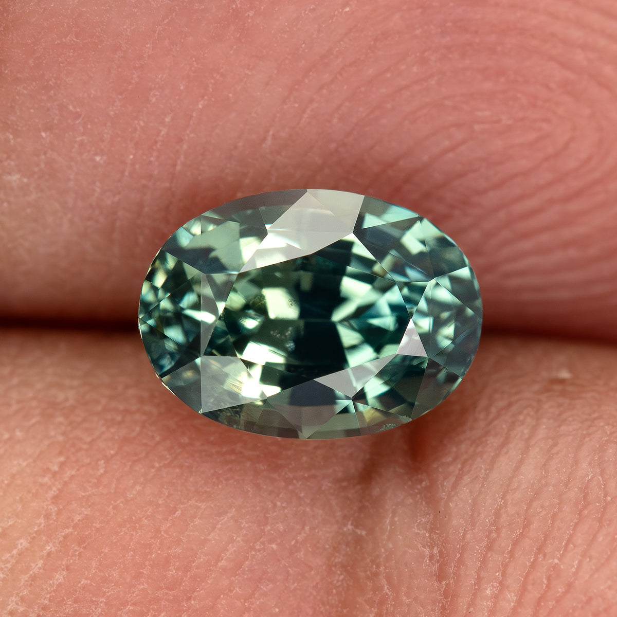 Teal-green oval sapphire resting face-up between fingertips, showing its complete outline at close range.