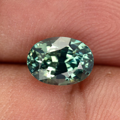 Teal-green oval sapphire resting face-up between fingertips, showing its complete outline at close range.