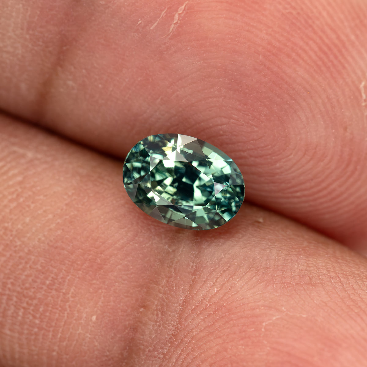 Wider view of the teal-green oval sapphire between fingertips, illustrating its physical scale.