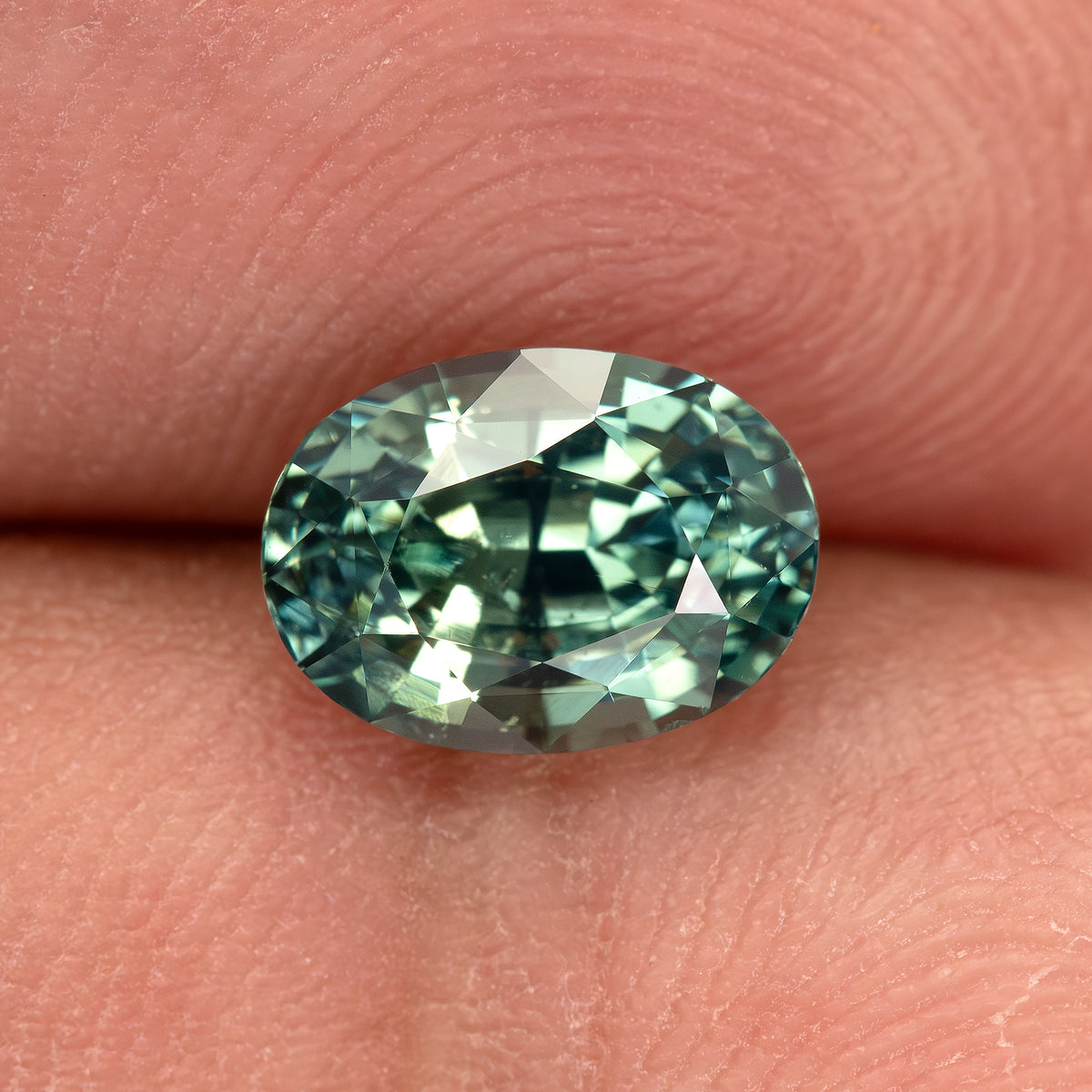 Teal-green oval sapphire on fingertips, with bright green and pale reflections across the crown.