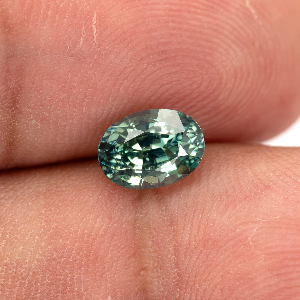 Teal-green oval sapphire resting at a low angle between fingertips, showing its crown height.