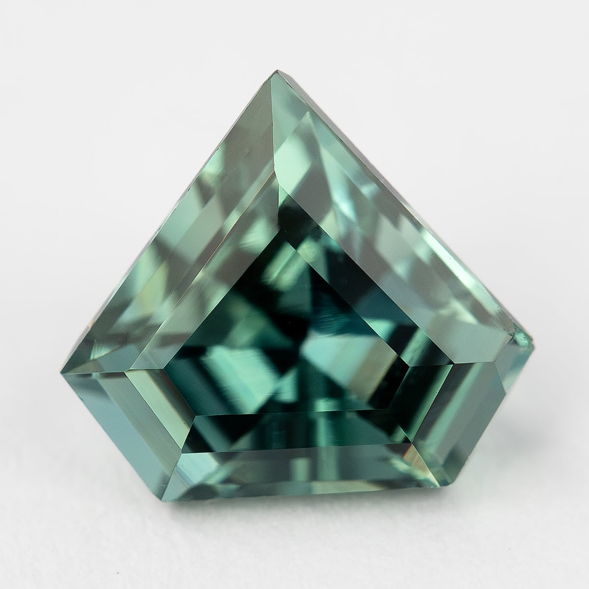 Alternate face-up view of the shield-cut sapphire on a light background, showing teal and blue-green reflections.
