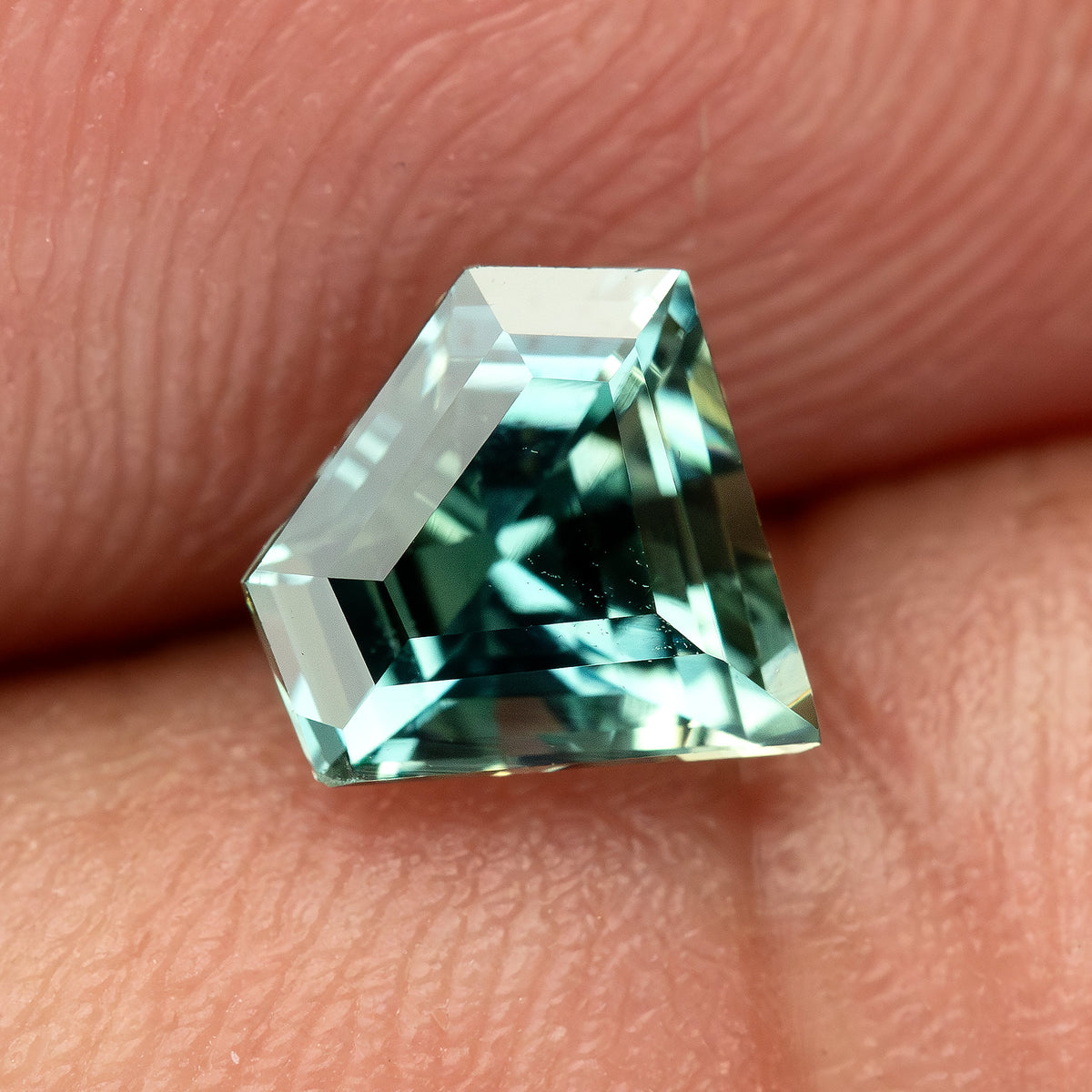 The teal-green shield sapphire resting on a fingertip, providing a visual scale reference.