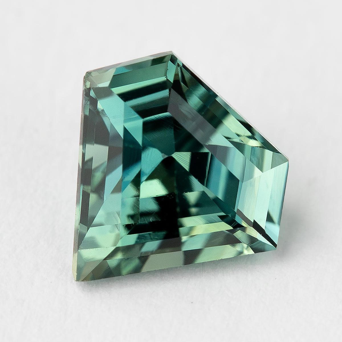 Tilted view of the sapphire on a light surface, showing the asymmetrical shield outline and stepped geometric reflections.