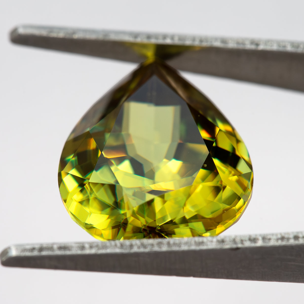 Pear-shaped sphene tilted in tweezers with a pale central reflection and green-yellow crown facets.
