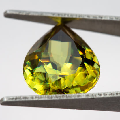 Pear-shaped sphene tilted in tweezers with a pale central reflection and green-yellow crown facets.