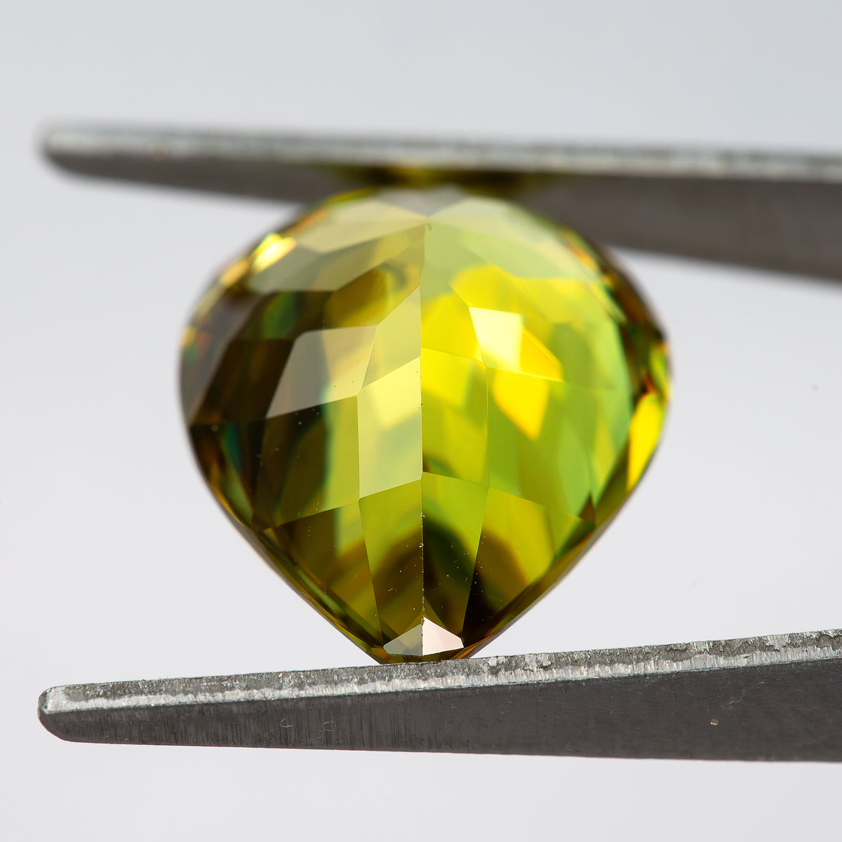 Angled pavilion-side view of the pear-shaped sphene in tweezers, showing bright yellow-green reflections.