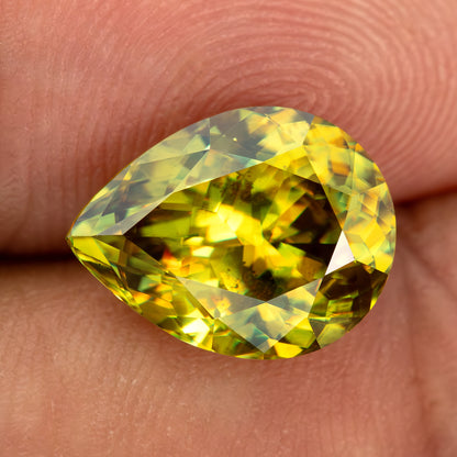 Green-yellow pear-shaped sphene resting face-up across fingertips, showing contrasting bright and dark facets.
