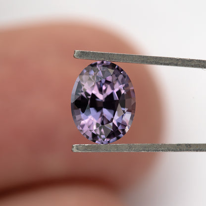 Oval spinel held face-up in tweezers against a blurred finger background.