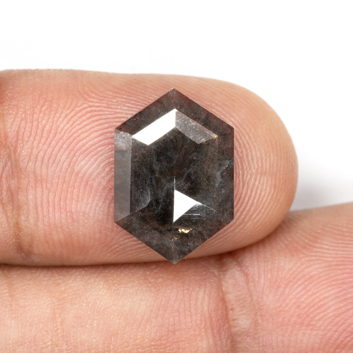 Hexagonal gray diamond resting on a fingertip at a slight angle, with dark inclusions and reflected facets visible.