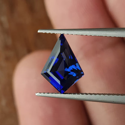 Royal blue lab-grown kite sapphire held face-up in tweezers, showing a long point, broad shoulders and geometric reflective facets.