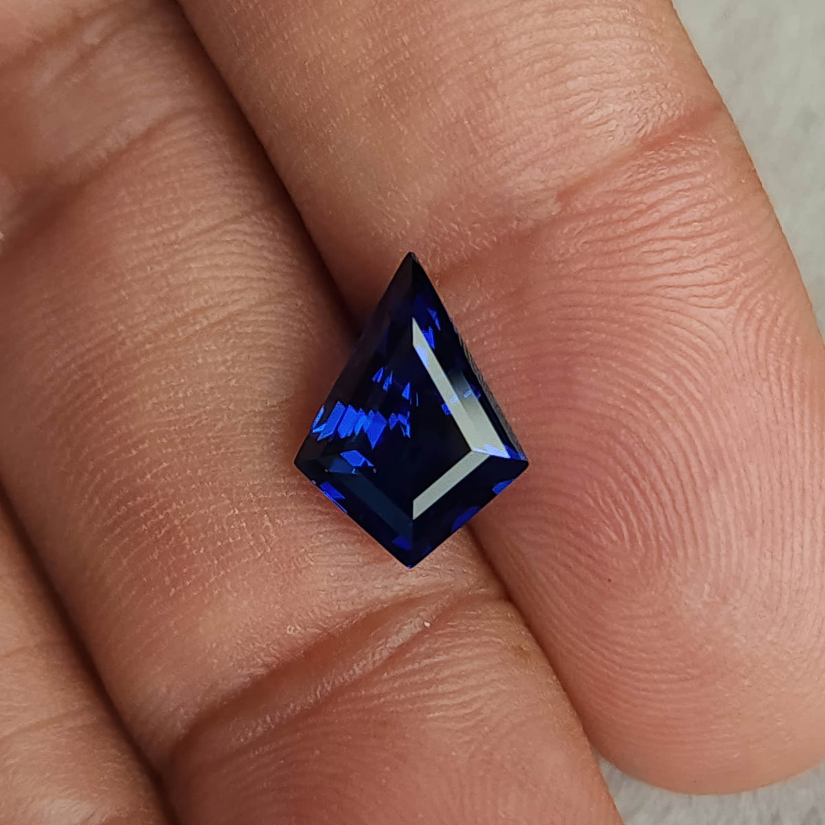 Blue kite-cut sapphire resting between fingers with its long point upward, showing scale and dark blue facet reflections.
