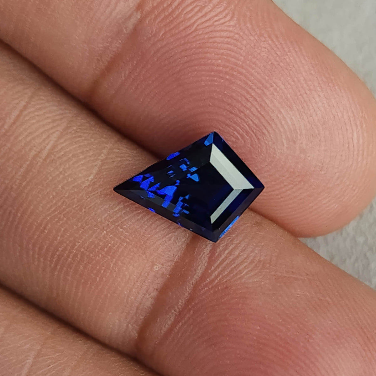 Royal blue kite sapphire lying sideways across fingertips, with a bright pale reflection along one edge.