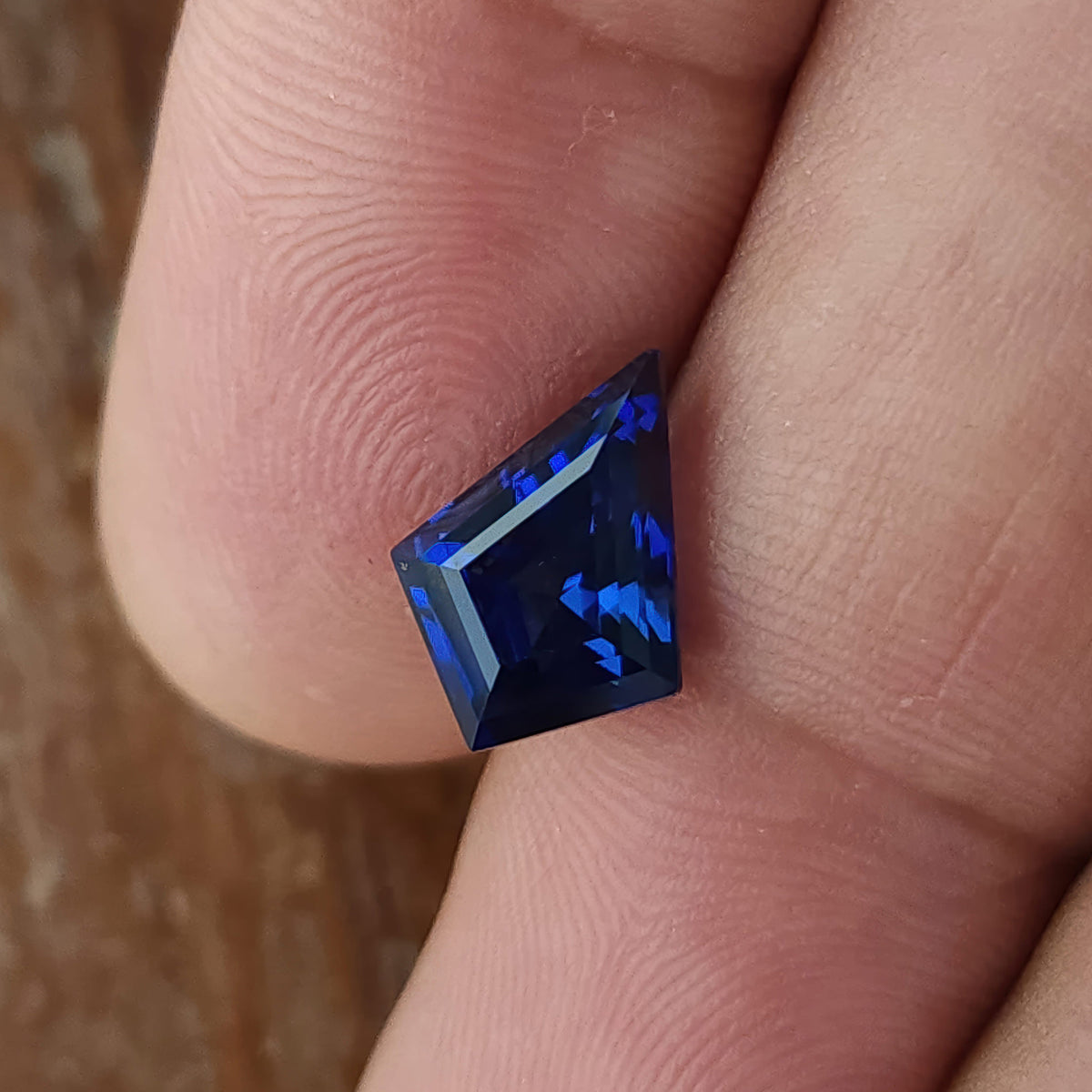 Kite sapphire held against a fingertip at an angle, showing blue flashes and a slanted pale facet reflection.